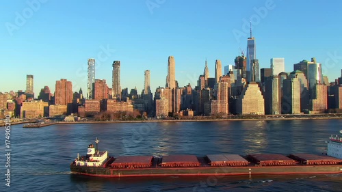 Wide, static, high-angle video of NYC Manhattan skyline with iconic skyscrapers. Golden hour light, clear blue sky. Calm East River foreground, cargo barge traversing. Concept of immense urban scale