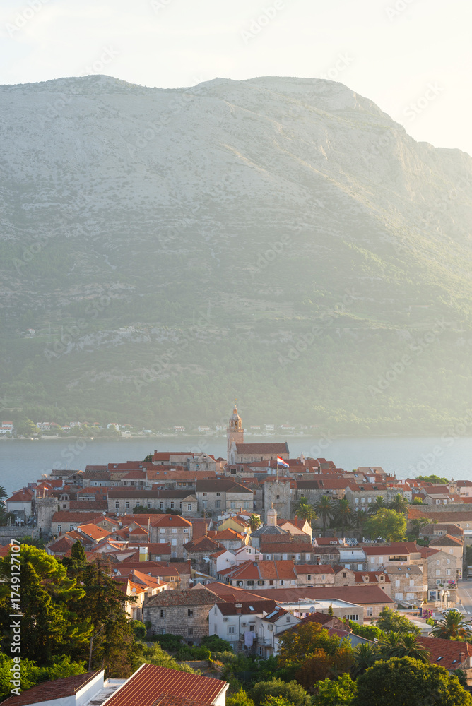 Fototapeta premium Die Altstadt von Korčula vor dem Gebirgsmasssiv des Sv. Ilija auf der Halbinsel Pelješac im Gegenlicht der Morgensonne, Insel Korčula, Süddalmatien, Kroatien