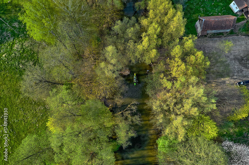Fototapeta Naklejka Na Ścianę i Meble -  Bird's-eye view of kayak paddling down brown river flanked by forest trees in Mazury, Poland on a sunny summer day