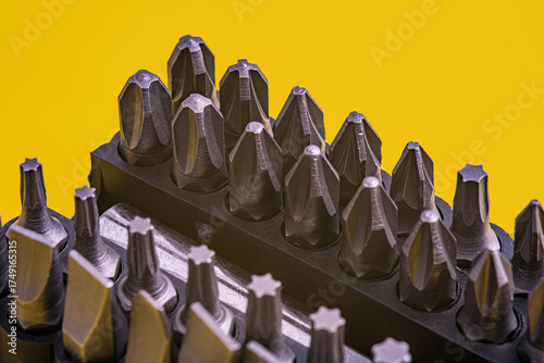 Close-up of screwdriver bit set with various metal tips arranged in a holder against a bright yellow background. Macro photograph of hand tools and craftsmanship. Selective focus.