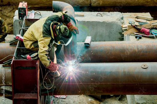 Obraz na plátně Man welder in full protective gear is welding a large metal pipe on a constructi