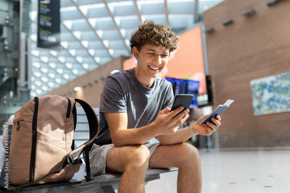 Obraz premium Smiling traveler guy sitting at airport bench using smartphone and holding passport. Happy young man waiting for boarding or booking hotel abroad