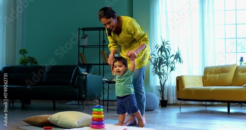 Indian infant boy walking holding young mother's hand in modern living room, learning to balance and take first steps with love, care, affection, and gentle guidance from mom