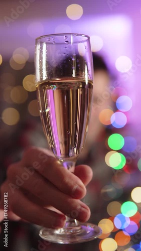 Vertical video close-up of male hand holding glass of champagne. Celebratory shot on New Years Eve featuring alcoholic sparkling drink in decorated illuminated setting.