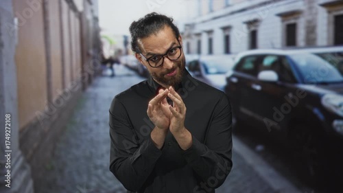 Man with glasses rubbing fingers together in black shirt on cobblestone street; scheming slyness cunning intent.