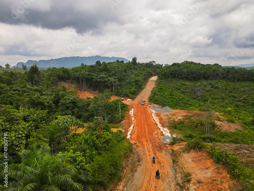 An aerial view of a muddy road under construction in the remote interior of East Kutai, Borneo, Indonesia. It cuts through rainforests and palm oil plantations with karst mountains on the horizon.