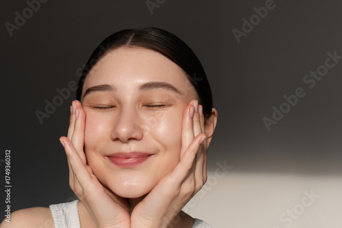 Asian woman with sleek dark hair smiling with closed eyes, gently cupping her glowing face, showing nourished, hydrated, and healthy skin in soft daylight. Skincare beauty routine portrait