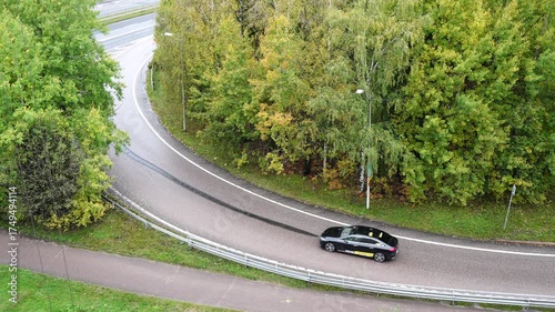Slow motion video of black car smoothly driving through winding forest road surrounded by lush green trees after rainfall, captured from above showing elegant motion and scenic countryside journey