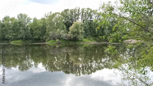 A view through the bushes onto the calm surface of a beautiful lake with dense bushes along the banks under a summer cloudy sky. Matveevskaya channel, Siberia, Russia.