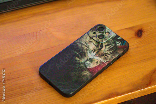 A smartphone lies on a wooden surface, featuring a custom case with a printed image of a sleeping tabby cat.