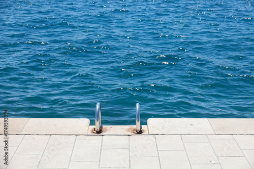 A metal ladder leading down to the swimming pool on the Zadar promenade, Croatia. Blue seawater and ripples in the background. Summer abstract reflections