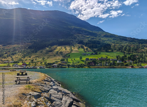 Scenic view of the turquoise water of Innvikfjorden with a picnic table on the rocky shore and green hills in Norway