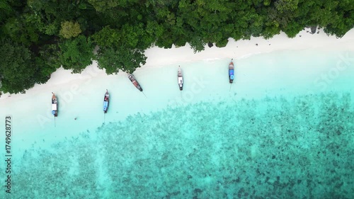 Aerial view of Monkey Beach on Ko Phi Phi Island, Thailand, showing turquoise sea, white sandy beach, tropical jungle and traditional long tail boats anchored near the shore.