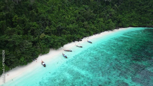 Aerial view of Monkey Beach on Ko Phi Phi Island, Thailand, showing turquoise sea, white sandy beach, tropical jungle and traditional long tail boats anchored near the shore.