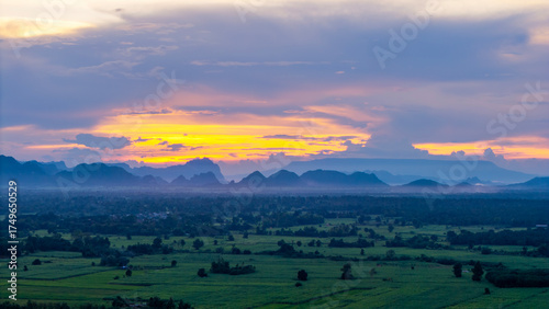 Breathtaking view of green farmland stretching toward silhouette of misty mountains under dramatic twilight sky, glowing in warm hues of orange yellow and purple.