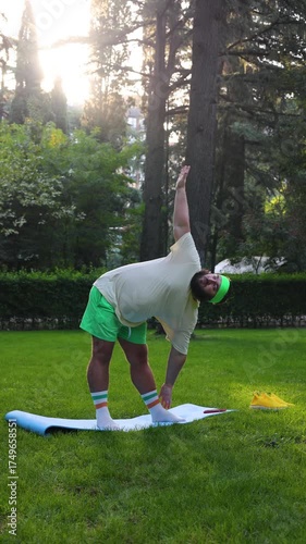 Vertical 4K street video. Fat man with beard, t-shirt, shorts, yellow sports sneakers man raises hand up, stands bend, triangle yoga pose, Trikonasana asana on blue mat on green grass in city park