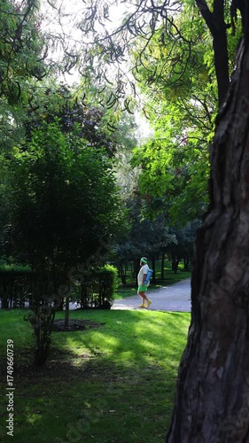 Vertical 4K street video. Wide view. Fat white man with beard, headband on head, green shorts, t-shirt, yellow sports sneakers, blue yoga mat walks on red treadmill in city park. Concept of warming up