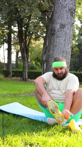 Vertical 4K street video. Fat white man with beard, green shorts, t-shirt sits on blue mat, takes off his yellow sport sneakers, watches yoga pose on mobile phone, video, read news, sms in city park