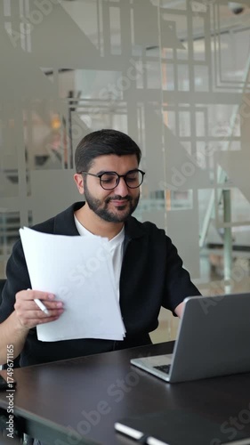 Male professional is seated at a desk in a contemporary office, examining important documents while a laptop and phone are nearby, deeply engaged in his task.