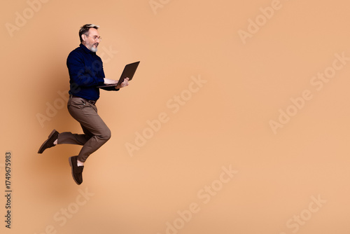 фотографія Energetic mature man in formal attire holding a laptop while jumping against bei