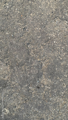 Detailed full-frame background texture of rough, damaged, dark gray asphalt pavement featuring embedded small gravel and vertical lines of erosion and cracking. Construction concept