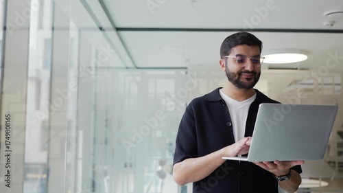 Business professional stands confidently in modern office holding laptop while contemplating next project or decision during work hours