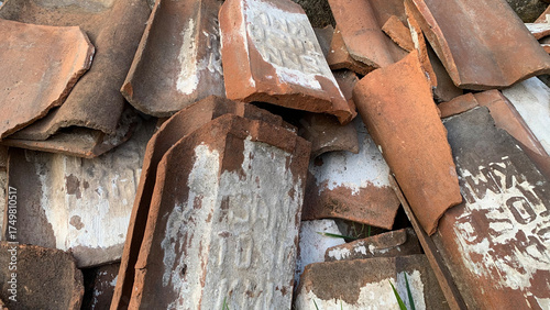 Detailed texture of an overlapping pile of broken, weathered terracotta clay roof tiles displaying remnants of white paint, aging, and faint stamped historic markings. Construction concept