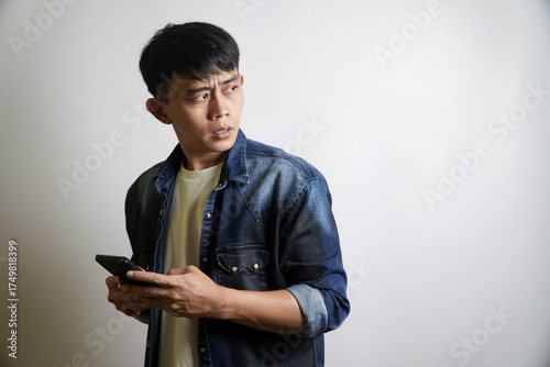 Billede på lærred Stunned Asian man holding a smartphone looks away with a shocked and fearful exp