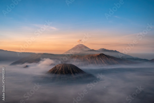 Mount Bromo volcano (Gunung Bromo) at sunrise, with a thin plume of smoke rising from its crater. The volcano is surrounded by a sea of mist, creating a mystical and serene atmosphere. Photographed fr