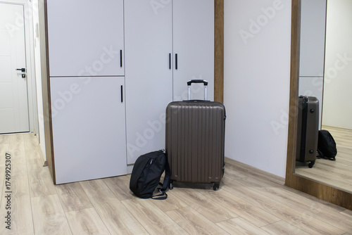 A bright apartment hallway: a rolling suitcase and backpack beside a wardrobe and tall mirror. Theme of arrival check out, short-term rental, business or leisure visit.