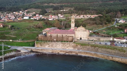 Aerial drone footage moving away from the Monastery of Santa María de Oia, showcasing its historic architecture, coastal setting, green fields, and waves crashing on the rocky shore  
