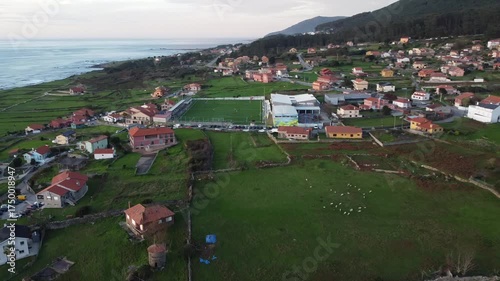 Aerial drone footage flying over Santa María de Oia, showcasing vibrant green fields, colorful houses, a coastal road, and the vast ocean stretching to the horizon  
