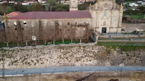 Aerial drone footage flying over the Monastery of Santa María de Oia, showcasing its historic stone architecture, red-tiled roofs, and detailed facade surrounded by green countryside  
