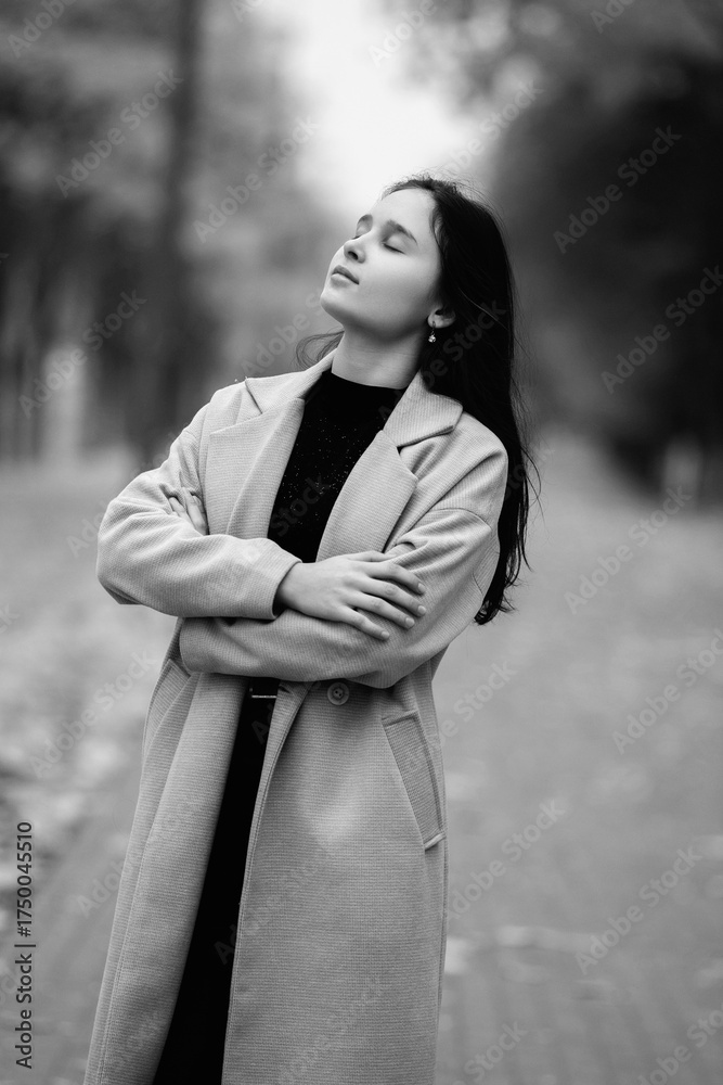 Fototapeta premium Asian woman in a stylish coat stands confidently on a tree-lined path, showcasing a serene expression and elegant posture, embodying grace and poise in a tranquil outdoor setting