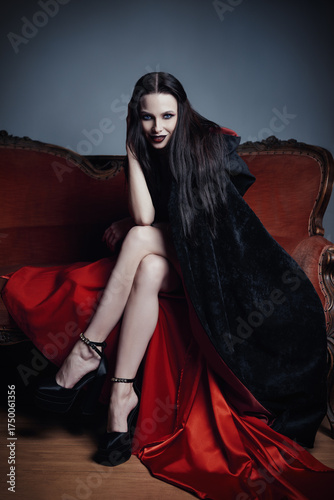 Dark beauty: seductive and dangerous young woman in image of witch sitting on sofa. Portrait of smiling vampire girl