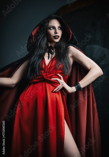 Sexy witch in red dress and black robe standing at the old mirror
