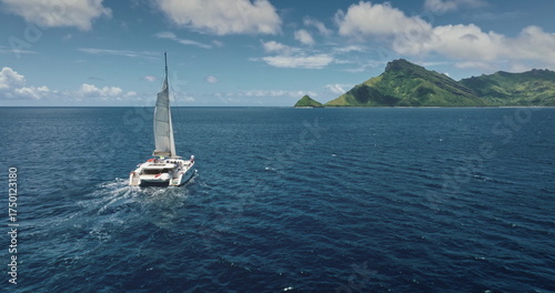 Fiji, Kuata island: White catamaran with tourists sailing blue ocean water near tropical island, under bright sunny sky. Remote wild nature paradise, exotic summer travel. Aerial drone flight