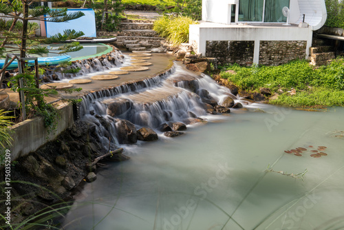 Water flows down over stone steps into a turquoise pond at De'camp hill ka-ang, Nakhon Nayok, Thailand. A tranquil scene of artificial waterfall in tropical-style garden resort.