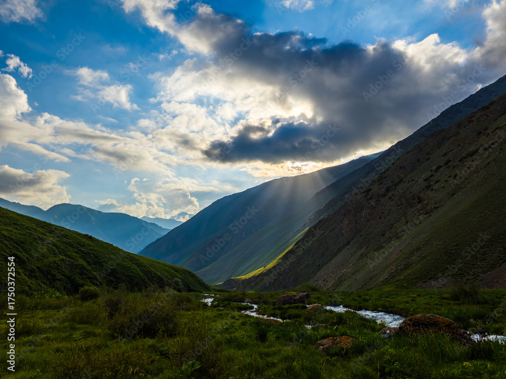Fototapeta premium Serene mountain valley scene showcases a river winding through majestic mountains. Sunlight breaks through clouds, creating a peaceful and awe-inspiring atmosphere.