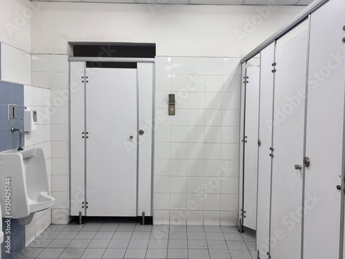 A simple, clean image of a public restroom showing a row of white stalls with metal hardware, a single urinal, and white tiled walls.