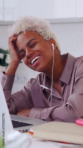 Young African American professional woman wearing earbuds, engaging with laptop and smiling during online meeting in a modern office, notebook and apple on desk; candid remote work emotion and focus