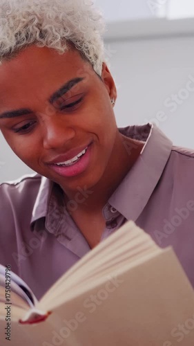 Portrait of a happy young woman with short curly hair reading a hardcover book indoors, smiling and turning pages, natural light illuminating her face, conveying relaxation, learning and quiet joy.