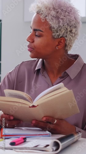 Solo female student or freelancer seated at a tidy workspace reading a paperback notebook, quietly mouthing words while preparing a presentation, attentive posture and casual clothing in an indoor