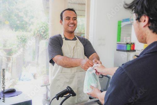 young asian man cashier worker standing near cashier counter in black apron and making payment and giving goods to customer in grocery store or modern market