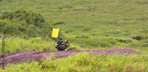 Camouflaged personnel skillfully crouches behind specialized equipment on a reddish-brown dirt rise, marked by a bright yellow flag, amidst a sprawling landscape of lush, wind-blown green grasses.