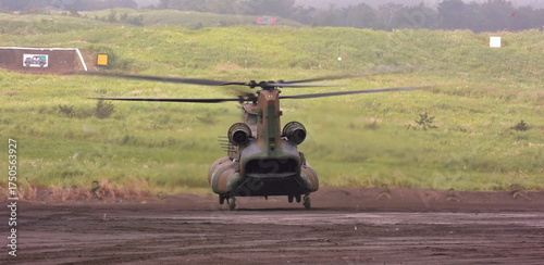 The robust tandem-rotor aircraft, painted in camouflage, dominates the muddy airstrip as it readies for flight against a backdrop of green, overcast hills and distant firing range targets.