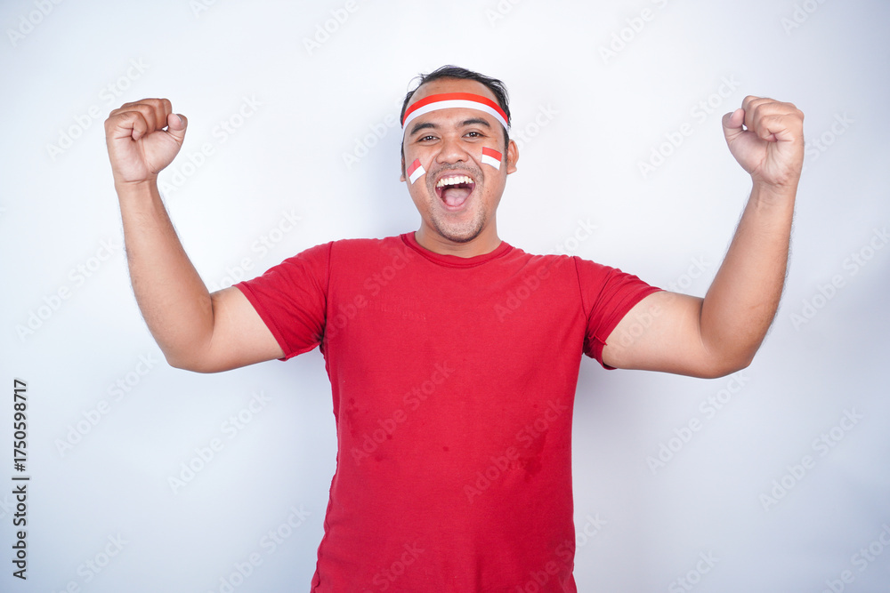Naklejka premium A young Asian man with a happy successful expression, raised a fist wearing red shirt and flag headband isolated by white background. Indonesia's independence day concept.