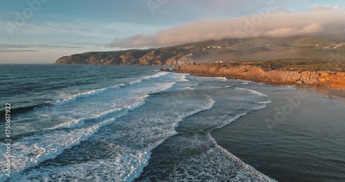 Portugal, Lisbon: Praia do Guincho coastline with white foam waves crashing on golden rocky cliffs under wide clear sky, remote coastal beach aerial landscape. Travel background. Drone flight footage