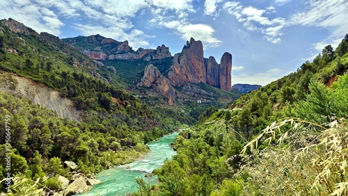 Mallos de Riglos in Spain features striking, vertical rock formations set against a backdrop of clear blue sky and scattered clouds. The towering red and brown cliffs, surrounded by dense green pine 