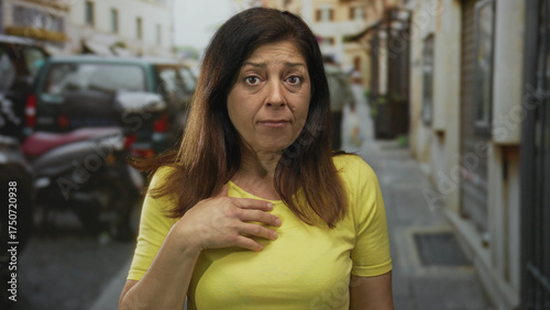 Free Download Middle aged woman in yellow shirt clutching chest near parked car on city street; pain concern heartattack. - GFXLoad